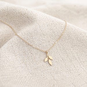 Peut inclure: Un délicat collier doré avec un pendentif en forme de feuille. Le pendentif comporte trois feuilles. Le collier est présenté sur un tissu texturé de couleur neutre.