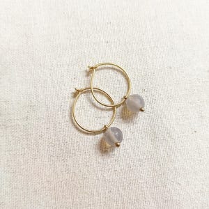 Puede incluir: Un par de pendientes de aro dorados, cada uno con una pequeña cuenta redonda de color gris pálido. Los pendientes se muestran sobre una superficie blanca texturizada. Diseño minimalista y elegante.