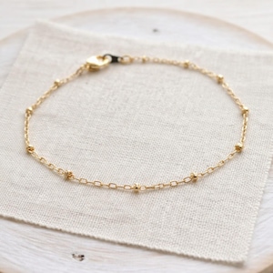 Könnte beinhalten: Zarter goldfarbener Fußkettchen mit kleinen, gleichmäßig verteilten Perlen und einem Kettenglied-Design. Das Fußkettchen wird auf einem weißen Leinentuch vor einem hellen Holzgrund präsentiert. Ein schlichtes und elegantes Schmuckstück.