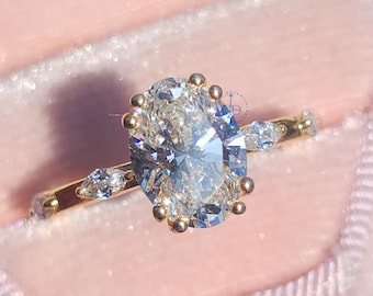 May include: A gold engagement ring with an oval-cut diamond center stone. The ring features smaller accent stones on the band and is set in a pink velvet ring box. The ring is a classic and elegant piece of jewelry.