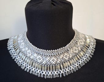 May include: A wide collar necklace crafted from intricate white and silver seed beads. The design features a geometric pattern with rows of metallic silver tube beads and a fringe of small, dangling crystal-like beads along the bottom edge.