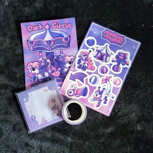Puede incluir: Un conjunto de artículos de papelería con temática de circo oscuro. Incluye un bloc de notas, una hoja de pegatinas y un rollo de cinta washi. El bloc de notas y la hoja de pegatinas presentan ilustraciones de artistas de circo y un tiovivo. La paleta de colores es morado, rosa y azul.