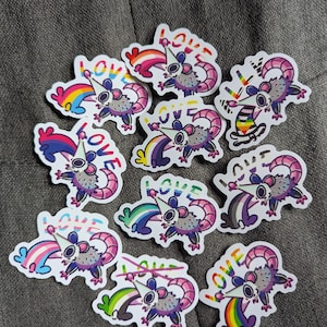 Puede incluir: Una colección de pegatinas coloridas con un dibujo de zarigüeya con una cola de arcoíris y la palabra "LOVE" en letras de arcoíris. Las pegatinas tienen varios diseños de banderas del orgullo, incluyendo banderas de arcoíris, transgénero y bisexuales.