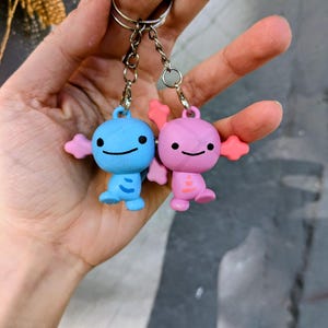 Può includere: Due piccoli portachiavi a forma di creature in stile cartone animato attaccati a un anello di metallo argentato. Una figura è azzurra con segni blu scuro, l'altra è rosa chiaro con segni arancioni. Entrambe hanno teste rotonde, occhi neri e sorrisi.