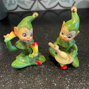 May include: Two vintage ceramic elf figurines. Each elf is dressed in a green outfit with a matching hat and red shoes. One elf holds a yellow and red object, while the other holds a wooden spoon and plate. Both have gold accents and are on a black surface.