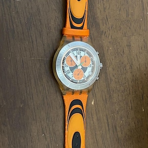 May include: Orange Swatch watch with a silver-colored face and orange strap. The watch face has black and orange details. The strap has black accents and a silver buckle. The watch is on a wooden surface.