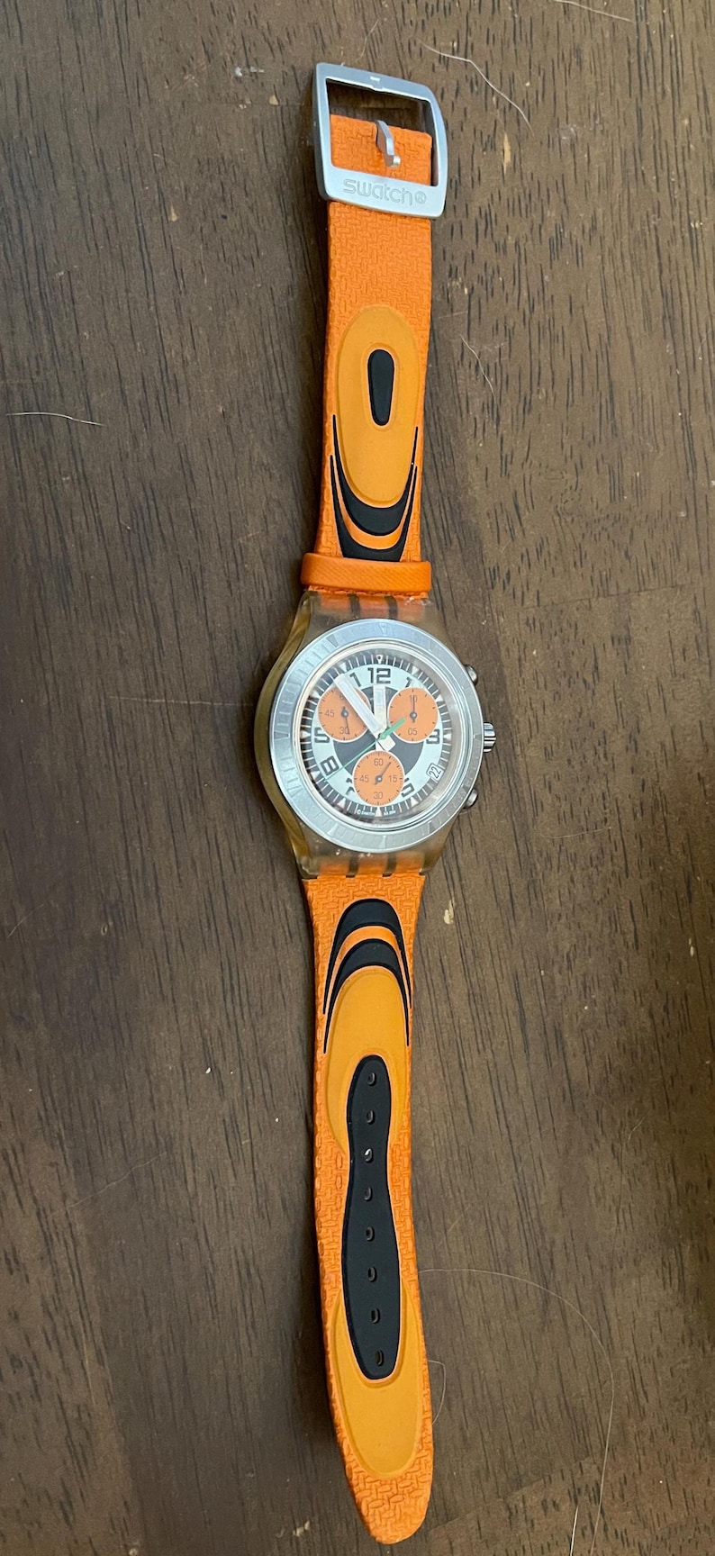 May include: Orange Swatch watch with a silver-colored face and orange strap. The watch face has black and orange details. The strap has black accents and a silver buckle. The watch is on a wooden surface.
