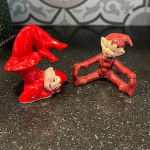 May include: Two red ceramic elf figurines. One is upside down with a white face and the other is sitting with arms and legs spread. The figurines have a glossy finish and are on a dark speckled surface.