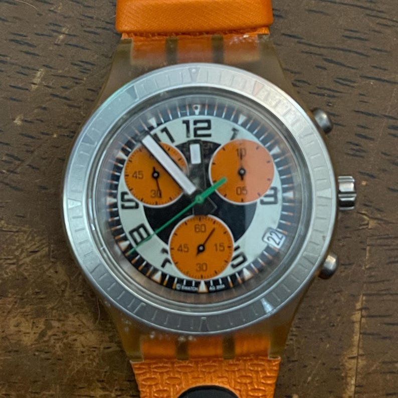 May include: A Swatch chronograph watch with an orange strap and a transparent case. The watch face features black, white, and orange details, including subdials and a date display. The watch has a silver bezel.