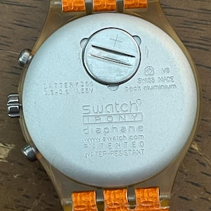 May include: Close-up of the back of a Swatch Irony Diaphane watch with an orange strap. The watch face is silver with the text "SWISS MADE" and "WATER-RESISTANT". The watch strap is orange.