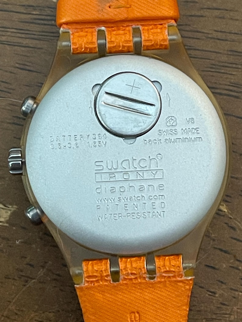 May include: Close-up of the back of a Swatch Irony Diaphane watch with an orange strap. The watch face is silver with the text "SWISS MADE" and "WATER-RESISTANT". The watch strap is orange.