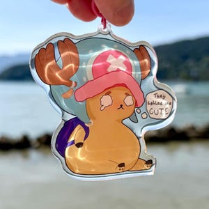 Op de afbeelding: Een heldere acryl bedel met een stripfiguur met bruine geweien, een roze hoed en een blauwe rugzak. Het personage is lichtoranje met een verdrietige uitdrukking. Een tekstballon zegt: "They called me CUTE".