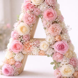 May include: A wooden letter "A" adorned with an array of pastel-colored flowers. The arrangement features pink, peach, and cream roses, along with delicate white filler flowers. The floral design creates a romantic and elegant aesthetic.