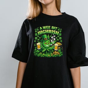 May include: Black t-shirt with a graphic featuring a cannabis leaf character wearing a green hat and smoking a cigarette. The text "A wee bit HIGHRISH" is above the character. The character is holding two mugs of beer and surrounded by shamrocks and gold coins.