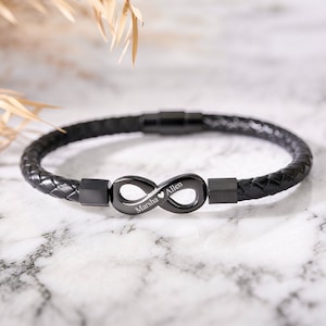 Puede incluir: Pulsera de cuero trenzado negro con un cierre de símbolo de infinito negro mate. El símbolo del infinito tiene grabados los nombres "Marsha Allen". Un accesorio elegante.