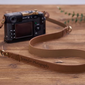 Op de afbeelding: Een bruinleren camerariem met de tekst "Jennifer Photography" ligt over een houten oppervlak. Op de achtergrond staat een zwarte camera met een gouden accent. De riem is gemaakt van lichtbruin leer.