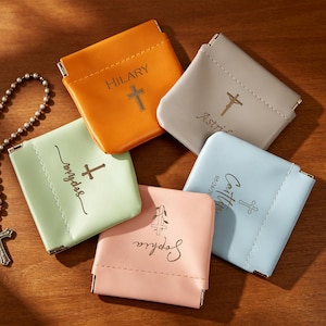 Puede incluir: Una variedad de monederos personalizados en varios colores pastel, cada uno con un nombre y un símbolo de cruz. Un monedero naranja dice "HILARY", uno gris "Astrid", uno azul claro "Caitlin", uno rosa "Sophia" y uno verde "Angelica". También hay un rosario con una cruz.