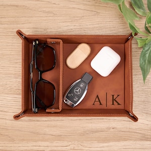 Op de afbeelding: Een bruin leren valet tray met compartimenten, met een zwarte zonnebril, een houten USB-stick, witte draadloze oortelefoons en een autosleutel. De tray is gepersonaliseerd met de initialen "A | K".