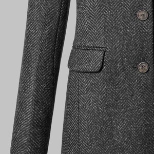 May include: Close-up of a dark gray herringbone tweed coat. The coat features a notched lapel, a single-breasted closure with three buttons, and a small pocket. The sleeve is visible, and the coat is tailored.