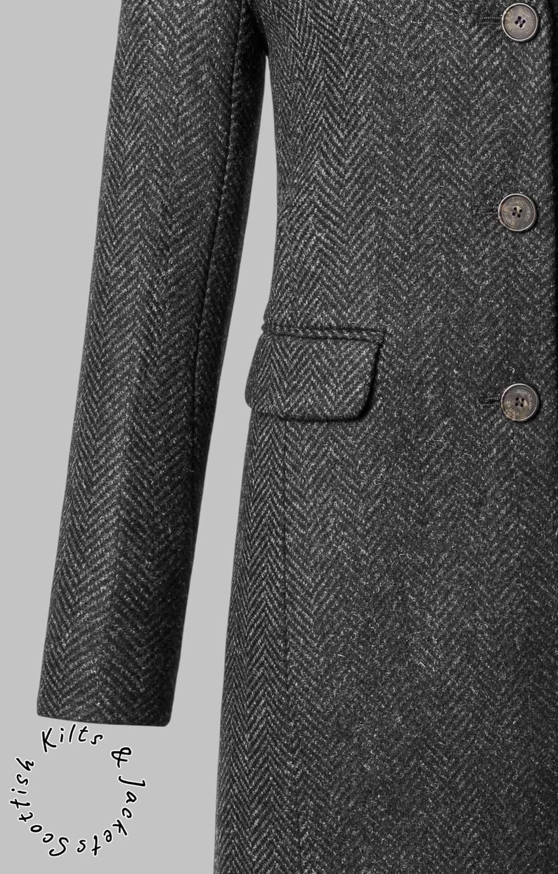 May include: Close-up of a dark gray herringbone tweed coat. The coat features a notched lapel, a single-breasted closure with three buttons, and a small pocket. The sleeve is visible, and the coat is tailored.