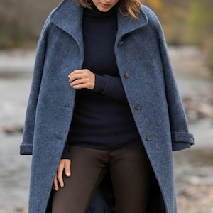 May include: A long, blue coat with a classic collar and button closure. The coat is worn over a navy blue sweater and brown pants. A small emblem is visible in the upper left corner. The coat appears to be made of a wool blend.