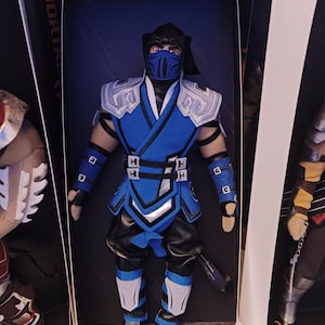 May include: A Sub-Zero action figure from the Mortal Kombat video game series. The figure is dressed in a blue and black outfit with silver accents, a mask, and a black hood. The figure is in a display box.