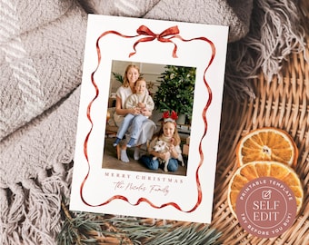 May include: A rectangular Christmas photo card featuring a watercolor red ribbon border with a bow at the top. The center displays a family photo with a Christmas tree. Text reads MERRY CHRISTMAS and The Nicolas Family in red lettering.