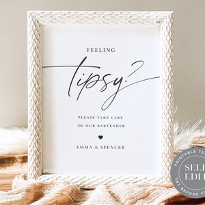 May include: White framed sign with black calligraphy that reads "Feeling Tipsy?" with the text "Please take care of our bartender" and "Emma & Spencer" below. The sign is on a wooden surface with a tan fabric in the foreground.
