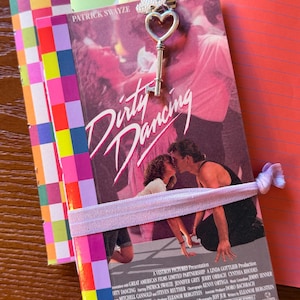 May include: A notebook featuring the Dirty Dancing movie poster on the cover, held closed by a light purple elastic band. A silver-toned metal clip with a heart-shaped key charm is attached to the top edge. The notebook is layered over other colorful paper.