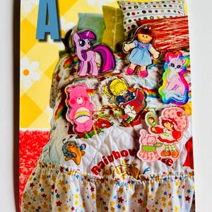May include: A colorful collage featuring various cartoon character stickers, including a pink My Little Pony, a doll, a Care Bear, and Strawberry Shortcake. The stickers are arranged on a bed with a rainbow quilt and a ruffled skirt with star patterns. The letter "A" is visible.