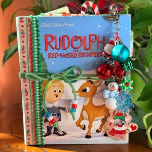 May include: A Little Golden Book titled Rudolph the Red-Nosed Reindeer, featuring a green suede tie, a decorative spine with red and green trim, and a metal charm dangle with miniature ornaments, including a snowman and a reindeer.