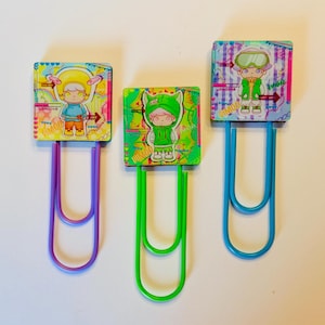 May include: Three decorative paper clips with colorful square designs and character illustrations. Each clip has a different color: purple, green, and blue. The designs feature cartoon figures in various outfits, with the word "DIMOO" visible on each clip.