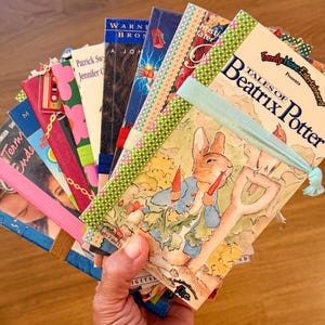 May include: A collection of VHS tapes arranged in a fan shape, featuring various movie covers including Tales of Beatrix Potter. Each tape is secured with a colorful elastic band and decorated with small charms, such as a cassette tape and gold chain.
