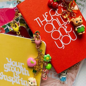 May include: A collection of colorful notebooks and charms. One red notebook reads "THERE'S ONLY ONE YOU" in white. A yellow notebook reads "Your growth is inspiring". Charms include a teddy bear, cherries, flowers, and a dog.