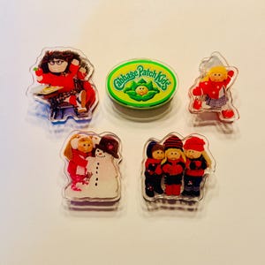 May include: A collection of Cabbage Patch Kids pins. The pins feature various characters in winter attire, a snowman, and the Cabbage Patch Kids logo. The pins are made of clear plastic with colorful graphics.