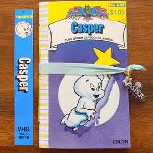 May include: A notebook featuring a Casper the Friendly Ghost VHS cover design with a yellow top section, a purple bottom section, and a light blue elastic band. A small charm with a white star and the word Casper hangs from the elastic band.