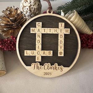 May include: A circular wooden Christmas ornament with a dark grey centre and a light wood frame. It features names spelled out with Scrabble tiles, the family name "The Clarkes", and the year 2025.