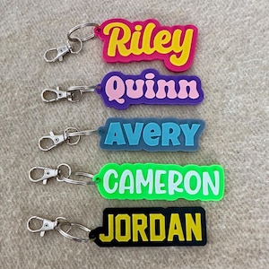 May include: A collection of colorful name keychains. Each keychain features a different name: Riley, Quinn, Avery, Cameron, and Jordan. The names are in bold, bubble letters with a contrasting border and attached to a silver clasp.
