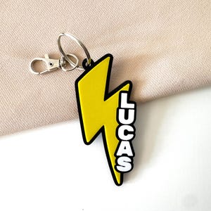 May include: A yellow lightning bolt keychain with the name "LUCAS" in white block letters. The keychain has a black outline and a silver clasp and keyring.