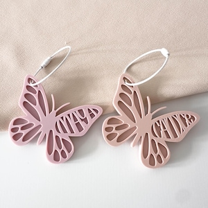 May include: Two butterfly-shaped luggage tags in pink and peach hues. Each tag features a name cut out of the butterfly's wing. They are attached to a white string with a metal clasp. The background is a light beige fabric.