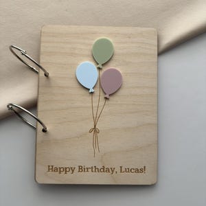 May include: A wooden photo album with a light wood grain finish. The cover features three pastel-colored balloons: green, blue, and pink. The text "Happy Birthday, Lucas!" is engraved below the balloons. The album is bound with silver metal rings.