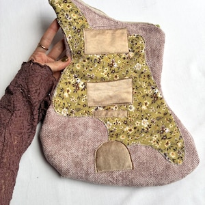 K&ouml;nnte beinhalten: Eine gitarrenf&ouml;rmige Rei&szlig;verschlusstasche aus mauvefarbenem Tweedstoff. Die Tasche hat ein Blumenmuster in Gr&uuml;n-, Wei&szlig;- und Braunt&ouml;nen. Beige rechteckige Aufn&auml;her sind auf den floralen Stoff gen&auml;ht. Der Rei&szlig;verschluss ist goldfarben.