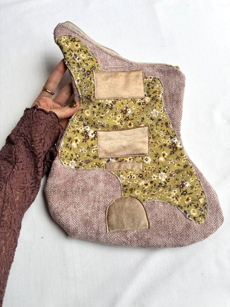 K&ouml;nnte beinhalten: Eine gitarrenf&ouml;rmige Rei&szlig;verschlusstasche aus mauvefarbenem Tweedstoff. Die Tasche hat ein Blumenmuster in Gr&uuml;n-, Wei&szlig;- und Braunt&ouml;nen. Beige rechteckige Aufn&auml;her sind auf den floralen Stoff gen&auml;ht. Der Rei&szlig;verschluss ist goldfarben.