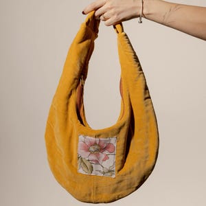 May include: Mustard yellow velvet shoulder bag with a floral patch. The bag has a curved shape, a zipper closure, and a matching mustard yellow strap. The floral patch features a pink flower with green leaves.