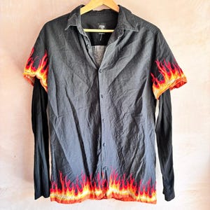 May include: A black button-down shirt featuring short sleeves with flame patterns in red, orange, and yellow. The hem also displays a matching flame design. The shirt includes long black sleeves layered underneath and a collar with a front button closure.