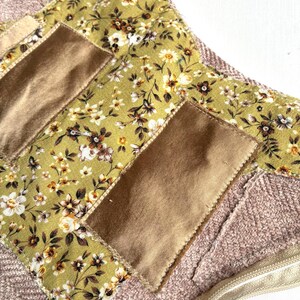 K&ouml;nnte beinhalten: Nahaufnahme eines handgefertigten Textilartikels mit einem floralen Muster in Gr&uuml;n, Braun und Wei&szlig;. Es hat rechteckige Patches in Beige und einen strukturierten rosa Hintergrund. Ein goldener Rei&szlig;verschluss ist sichtbar.
