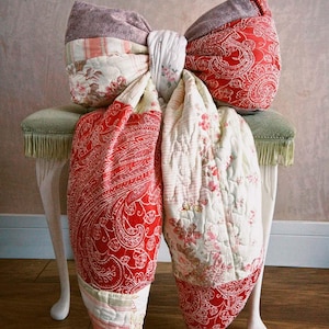 May include: A large decorative bow crafted from quilted fabric patches in red, white, and grey. The bow features a mix of paisley, floral, and herringbone patterns with two long, hanging fabric tails draped over a small, light-coloured stool.