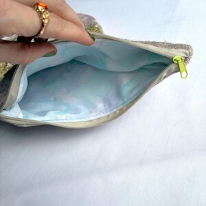 K&ouml;nnte beinhalten: Ge&ouml;ffnete, gezippte Tasche mit einem hellgr&uuml;nen Rei&szlig;verschluss. Die Au&szlig;enseite ist strukturiert und neutralfarben, w&auml;hrend die Innenseite ein hellblaues Blumenmuster aufweist. Die Tasche wird offen gehalten und zeigt das Innere.