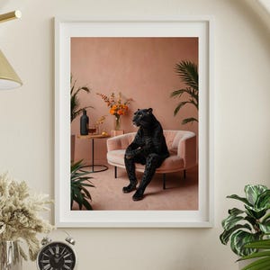 May include: A framed wall art print featuring a black panther sitting upright on a light pink velvet armchair. The scene includes a small round side table with a black bottle and a glass, a vase of orange flowers, and green potted plants.