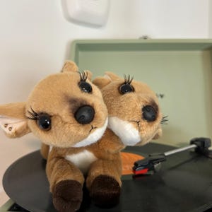 May include: A plush toy deer with two heads, brown fur, and large black eyes with long eyelashes. The deer is sitting on a black vinyl record on a green record player.